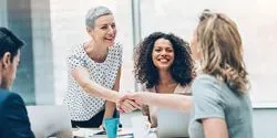Woman with Short Hair Shakes Hand of Blonde Woman While Dark-Haired Woman Looks On Smiling