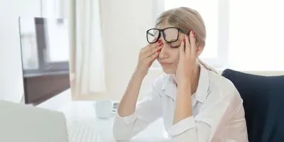 A stressed blonde female worker sits at her desk and rubs her eyes in exhaustion.