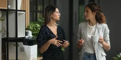 Two women speaking in an office setting. One woman is dressed in black the other in an off-white blazer.