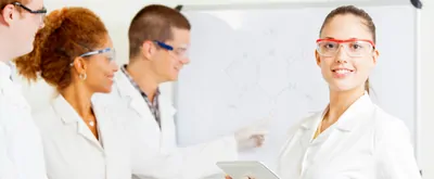 A young female scientist stands with a clipboard while three of her colleagues work on a whiteboard in the background. Everyone is wearing lab coats.