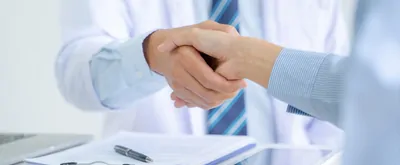 A closeup of a handshake between a lab-coated scientist and a businessperson in a dress shirt over top of a desk with a clipboard and tablet to illustrate the choosing of a CRO.