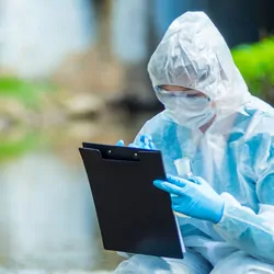 A scientist with a clipboard crouches down in a source of water, taking notes