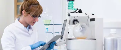 Chemical scientist wearing rubber gloves controls CBD oil extraction in rotavapor. Machine has big green condenser. Glass tubes and cups with hemp seed are part of laboratory background. Hero image.