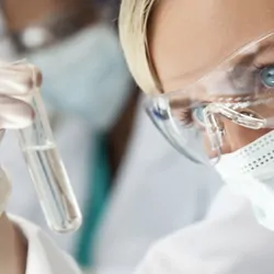 A photo of a woman in a cancer research lab wearing goggles and holding a test tube.