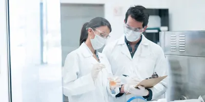 Two scientists wearing lab coats and masks fill out forms on a clipboard in the lab.