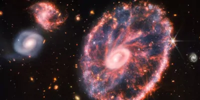 A large pink, speckled galaxy resembling a wheel with with a small, inner oval, with dusty blue in between on the right, with two smaller spiral galaxies about the same size to the left against a black background