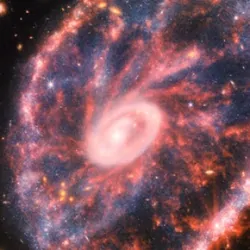 A large pink, speckled galaxy resembling a wheel with with a small, inner oval, with dusty blue in between on the right, with two smaller spiral galaxies about the same size to the left against a black background