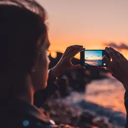 Woman taking photo of the ocean on her phone, at sunset