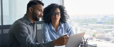 Indian male ceo executive manager mentor giving consultation on financial operations to female African American colleague intern using laptop sitting in modern office near panoramic window
