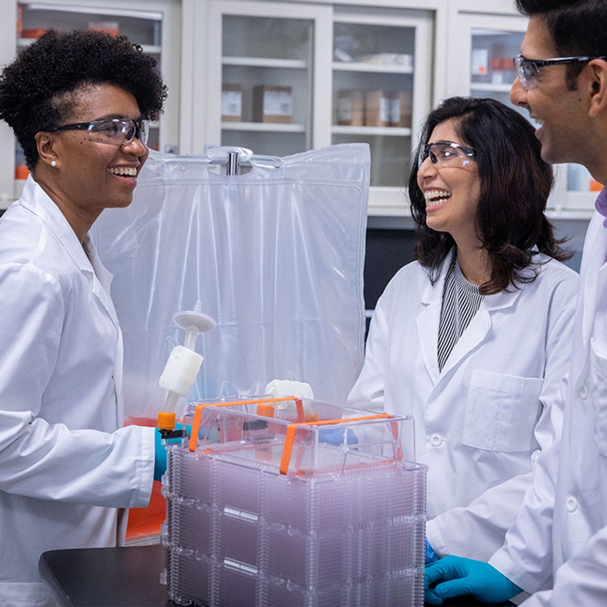 Three scientists are laughing together and setting up a HYPERstack cell culture vessel with large bags of cell culture media for large-scale culturing