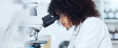 One mixed race scientist with curly hair wearing safety goggles and gloves analysing medical test samples on a microscope in a lab.