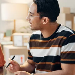 a man with tan skin and dark hair wearing a brown striped shirt talks to a brown skinned woman with black hair in an orange shirt while she is looking at a clipboard on a desk, in an office setting
