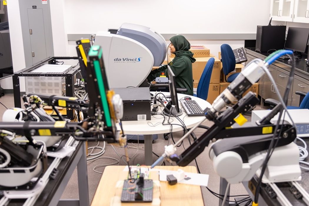 Researcher sits at a large gray machine with a blue-gray hood. Electronics equipment are scattered around the lab. Researcher sits at a large gray machine with a blue-gray hood. Electronics equipment are scattered around the lab.