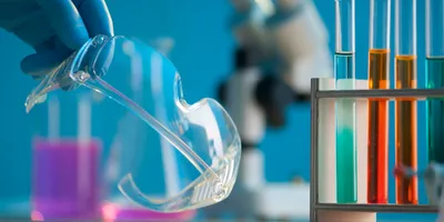 Close up of the gloved hand of alab worker grabbing goggles from a bench beside test tubes of coloured liquid