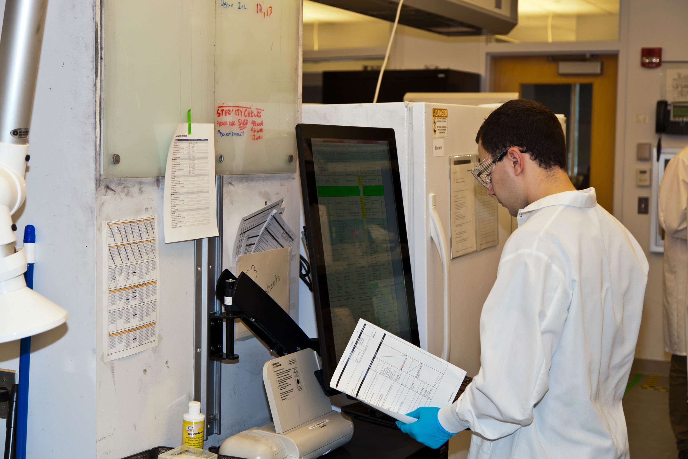 A male scientist with short hair and glasses holds a printed sheet of paper beside a machine with a touch screen