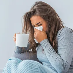 A sick woman sits on her bed holding a cup of tea and blows her nose