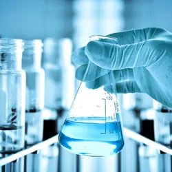 a gloved hand holding a flask of blue liquid in a chemistry lab suggesting the concept of batcha nd continuous flow chemistry