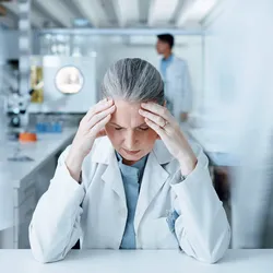 A stressed lab manager sits at her desk and rubs her temples as people walk behind her