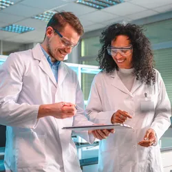 Two scientists in lab coats and safety goggles smiling and collaborating in a laboratory, fostering a positive and productive work environment.