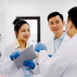 Three scientists in a laboratory discussing research, with one holding a tablet and another handling a test sample. The collaborative setting highlights teamwork, but understaffing in labs can impact efficiency and innovation