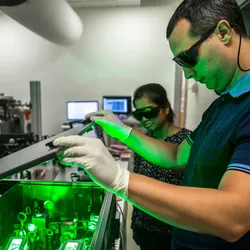 Two researchers wearing laser safety goggles and gloves work in a laboratory with high-powered green lasers; one is lifting the cover of a laser apparatus while green light illuminates the space—highlighting the importance of laser safety in lab environments.