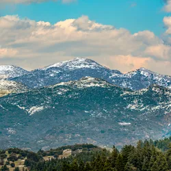 Snow-covered peaks of Mount Helmos in Greece, a research site for studying biological particles in clouds, with forested slopes beneath a partly cloudy sky.