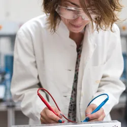 A female lab technician wearing a lab coat and safety glasses works on soldering a circuit board taken from a piece of lab equipment
