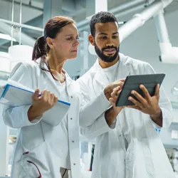 Two scientists stand in the lab discussing what's displayed on a tablet computer
