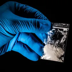 Handling fentanyl under proper fentanyl safety guidelines with gloved hand, and closed plastic bag of white, powdered fentanyl.