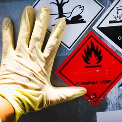 Gloved hand upon a container with multiple hazard signs on it.