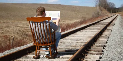 man sitting in a rocking chair reading a newspaper on railway tracks an example of male itiot theory
