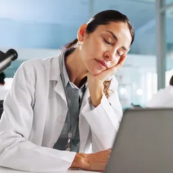 A laboratory professional wearing a white lab coat appears fatigued while working at a computer. In the background, a colleague focuses on research under a microscope, highlighting the demanding environment of modern laboratories. This image illustrates the importance of healthy sleep habits for lab staff to enhance focus, safety, and well-being.
