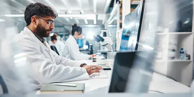 Two scientists, male and female sit at a bench. He is typing on a laptop, she is sitting in front of a microscope
