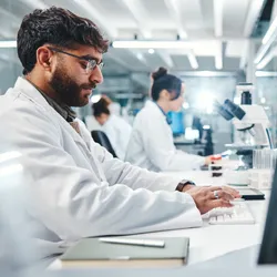 Two scientists, male and female sit at a bench. He is typing on a laptop, she is sitting in front of a microscope