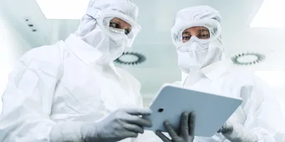 Two lab workers in full PPE looking at a tablet, symbolizing the monitoring of an environmental cleanroom.
