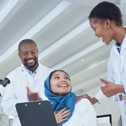 Three lab professionals smiling and engaging positively with one another in a bright laboratory, with one person seated holding a clipboard and two colleagues offering gestures of support and encouragement, surrounded by microscopes and lab equipment.