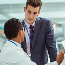 Two professionals in a laboratory setting discussing documents, illustrating collaboration and strategy as part of how to craft a persuasive business case that wins executive approval.
