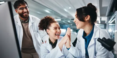 Three scientist in the lab; two of them are giving each other a high five while the third smiles, demonstrating the power of humor in the workplace