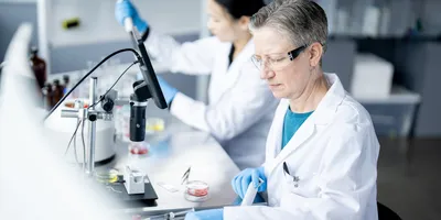 Gloved scientist inspects petri dishes while colleague pipettes—lab scene emphasizes traceability and data integrity.