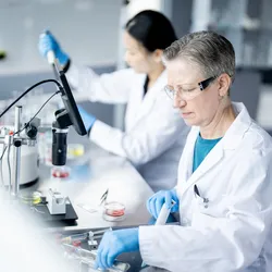 Gloved scientist inspects petri dishes while colleague pipettes—lab scene emphasizes traceability and data integrity.