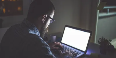 A man works late at night in a dimly lit room, illuminated by the glow of his laptop screen. He appears focused but fatigued, symbolizing the “infinite workday” described in Microsoft’s research. 