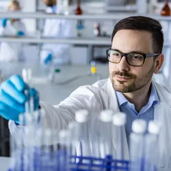Scientist places a test tube into a rack. Another scientist sits at a bench in the background