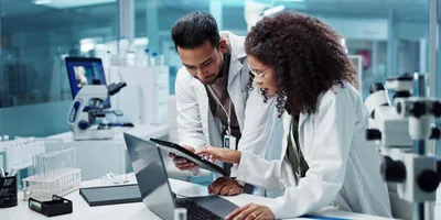Two scientists in a pharmaceutical lab reviewing data on a digital tablet and laptop surrounded by laboratory equipment, including microscopes and test tubes.
