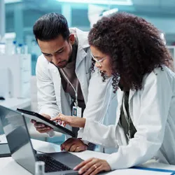 Two scientists in a pharmaceutical lab reviewing data on a digital tablet and laptop surrounded by laboratory equipment, including microscopes and test tubes.