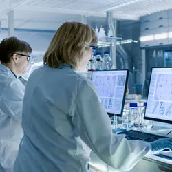 Scientists working in a pharmaceutical lab analyzing data on desktop monitors, with DNA sequences and molecular visuals displayed on screen.