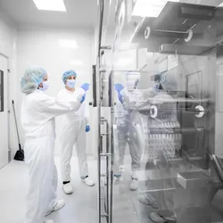 Pharmaceutical personnel in full cleanroom attire inspecting equipment inside a sterile, controlled environment with reflective surfaces and HEPA-filtered enclosures.