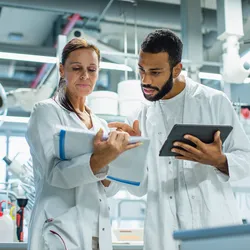 Two pharmaceutical scientists in lab coats discussing technical data, one holding a clipboard and the other using a tablet, in a modern laboratory setting.