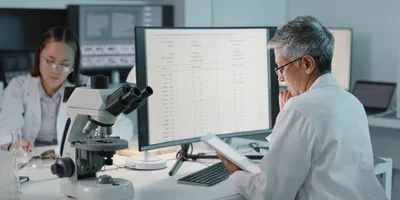 Two scientists in a laboratory analyzing test data on a monitor and tablet, with a microscope in the foreground, highlighting lab data integrity and digital systems.