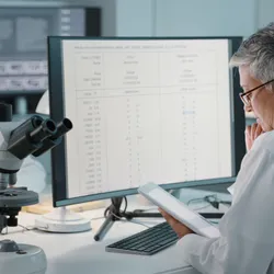 Two scientists in a laboratory analyzing test data on a monitor and tablet, with a microscope in the foreground, highlighting lab data integrity and digital systems.