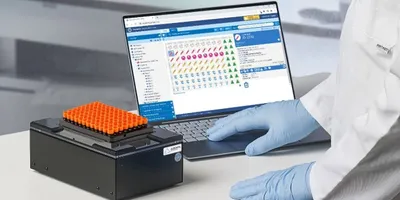 Scientist wearing lab gloves operating a sample tracking device connected to laboratory software on a laptop, with a 2D barcoded tube rack placed on the scanner in a clinical or research lab environment.